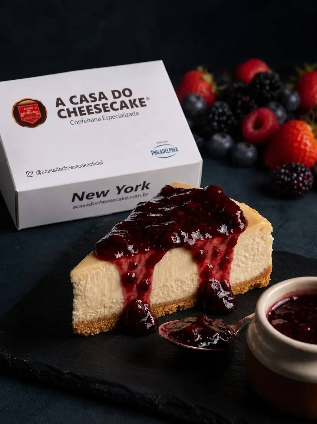 Fatia de New York Cheesecake com calda de frutas vermelhas em frente à caixa da Casa do Cheesecake