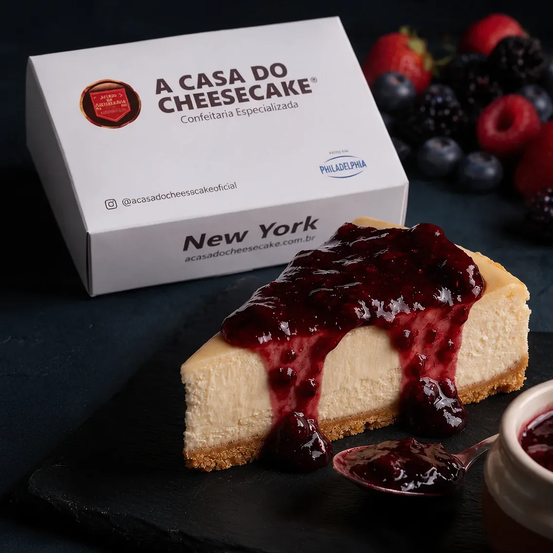Fatia de New York Cheesecake com calda de frutas vermelhas em frente à caixa da Casa do Cheesecake