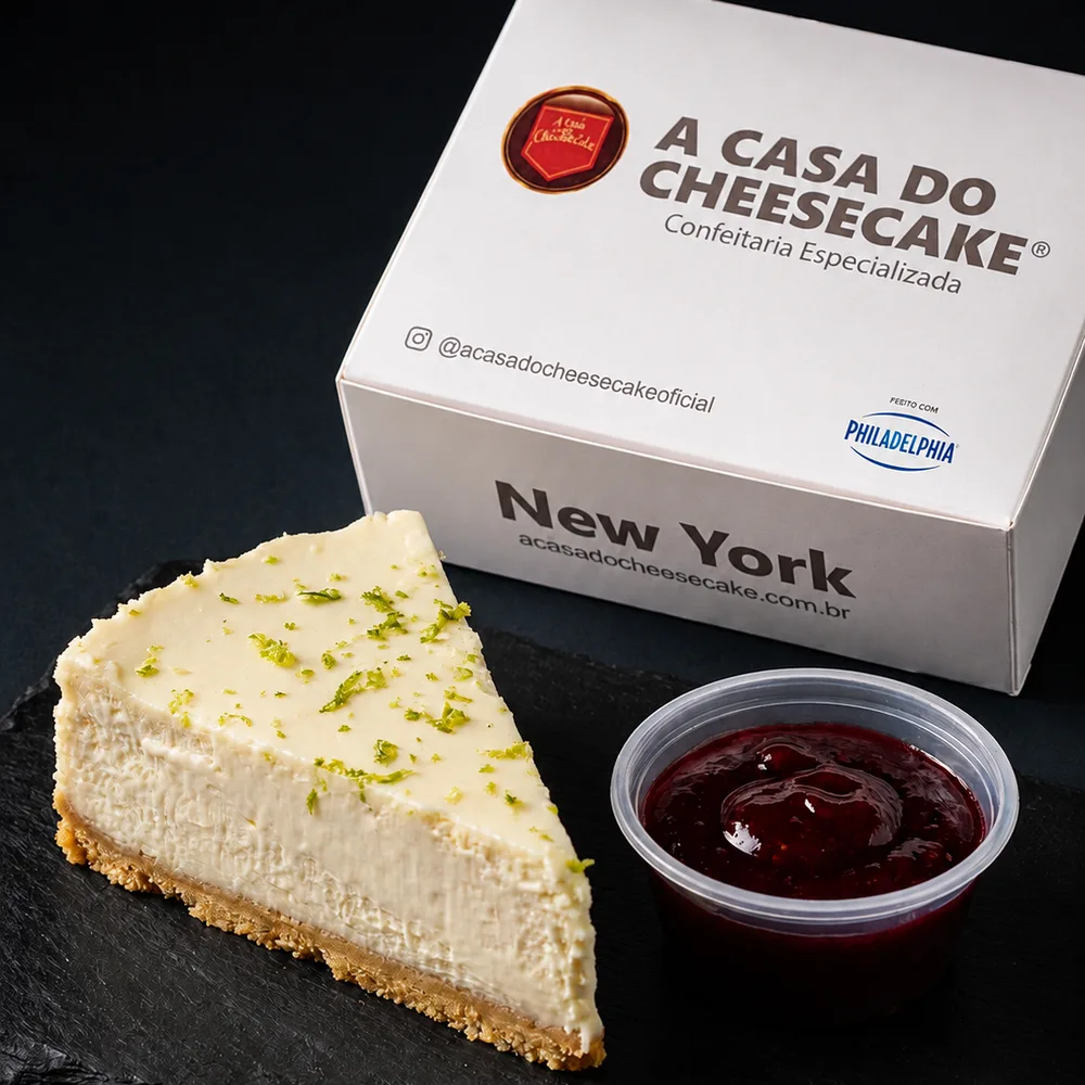 Fatia de cheesecake Limone com raspas de limão, ao lado da calda de morango e da caixa