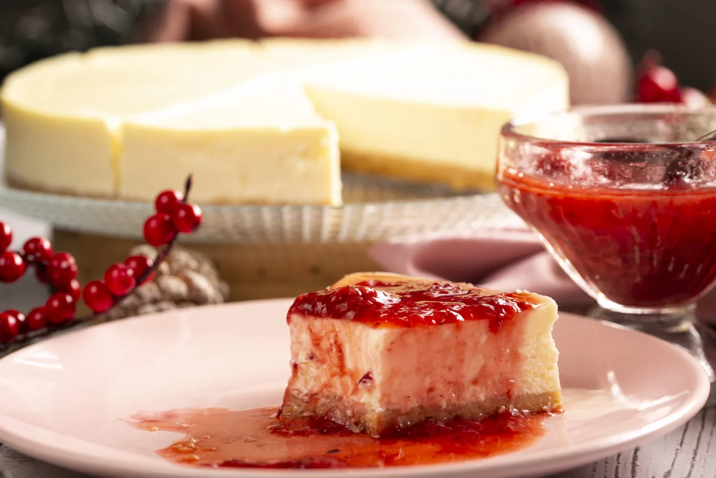 Fatia de cheesecake com calda de morango num prato, com o cheesecake inteiro ao fundo